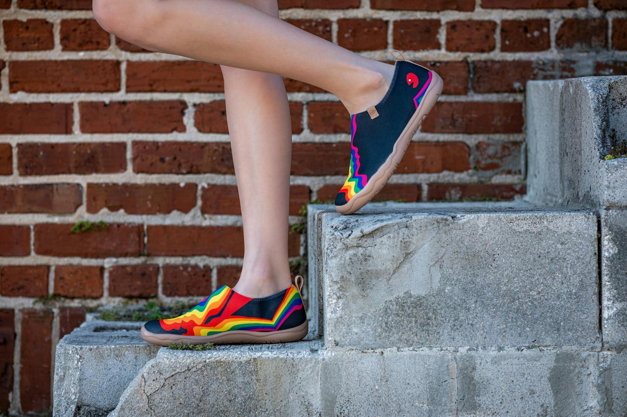 Vibrant rainbow sneakers worn on steps against a brick wall outdoors.